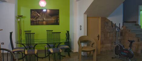 A living room with a green wall, a dining table with chairs, a wicker chair, a treadmill, and a staircase.