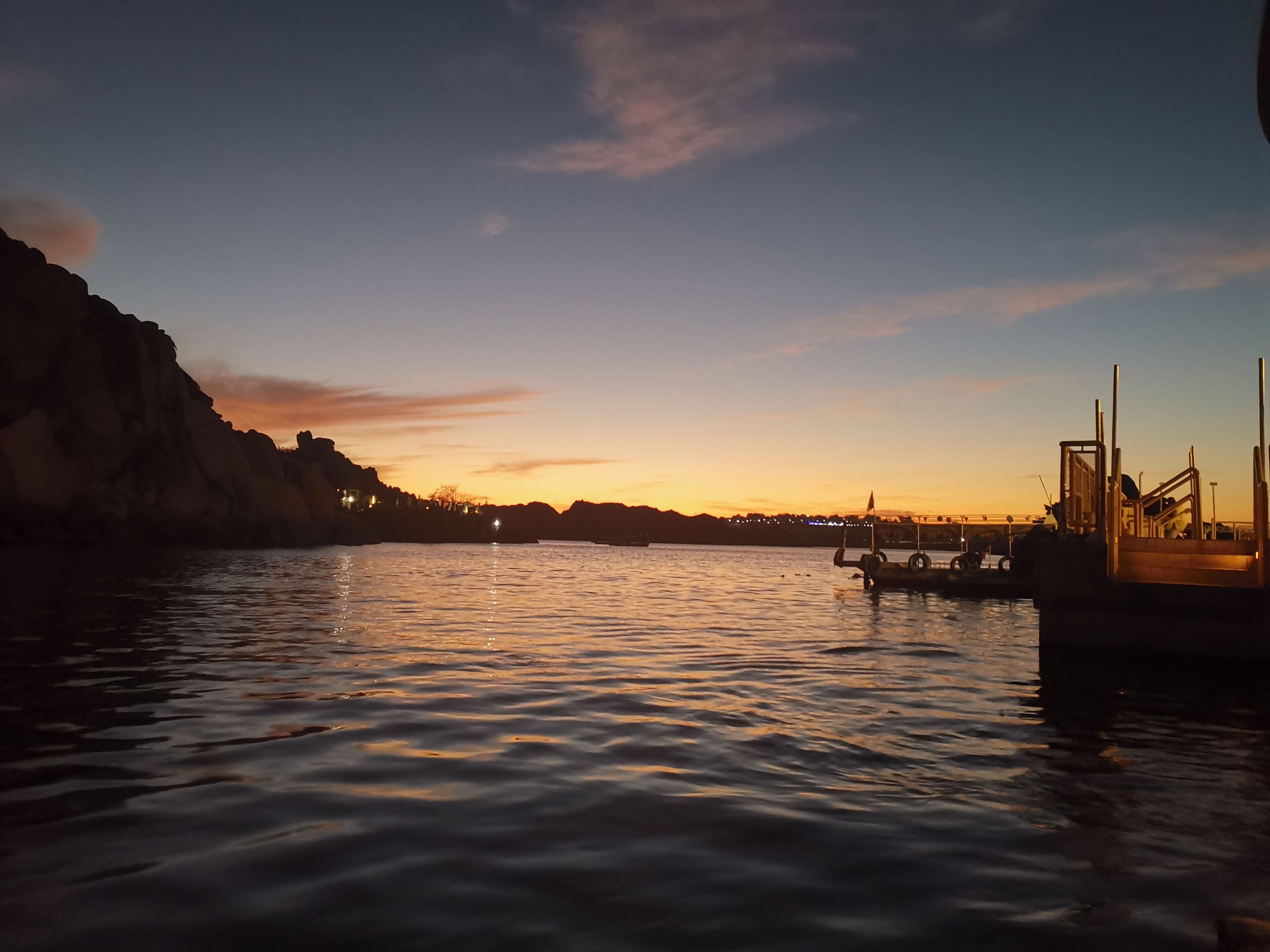 Una orilla tranquila al atardecer con barcos amarrados, un muelle y edificios a la distancia.