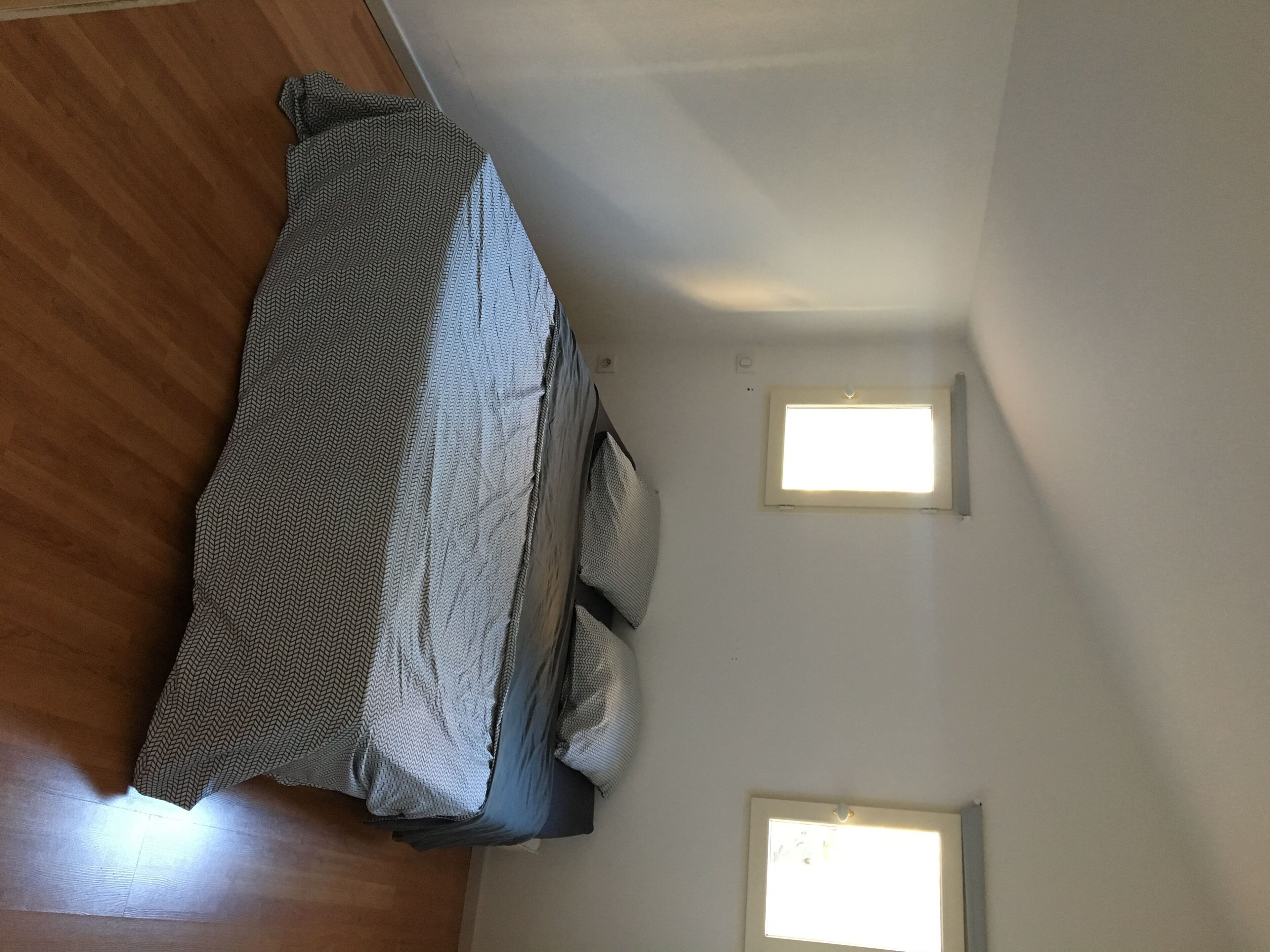 A bedroom with a bed covered in a patterned blanket, two bedside tables, and two wall-mounted windows.