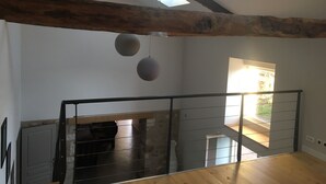 Una habitación amplia con techos altos, vigas de madera a la vista y tragaluces. Una escalera conduce a un nivel superior, y una puerta se abre a un espacio exterior luminoso.