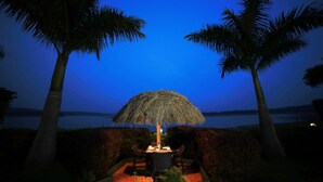 Un espace de restauration en plein air avec une table et des chaises sous un parasol en chaume, entouré de palmiers et offrant une vue sur l’eau.