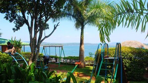 Un terrain de jeux avec des balançoires et un toboggan, entouré d’une végétation luxuriante et offrant une vue sur la mer.