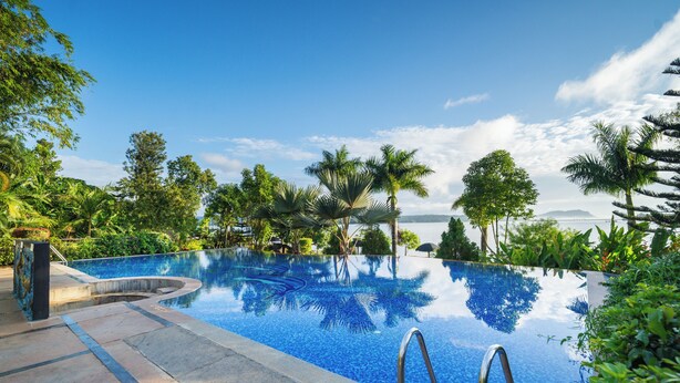 Une magnifique piscine à débordement d’un bleu profond, entourée d’une végétation luxuriante et de palmiers, avec une vue imprenable sur la mer et les montagnes en arrière-plan.