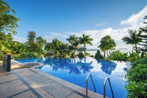 Una piscina infinita de aguas cristalinas rodeada de exuberante vegetación y palmeras, con vistas al mar y a las montañas al fondo.