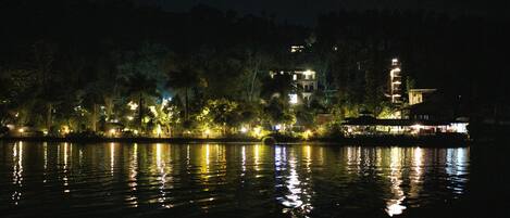 Vista nocturna de una propiedad junto al lago, con edificios e iluminacion en los árboles, reflejándose en el agua.