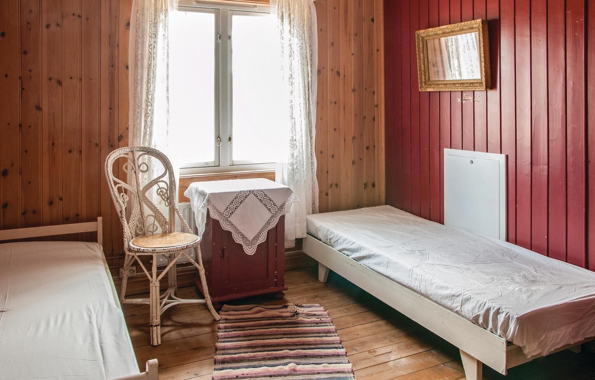 A small, cozy room with wooden walls, a single bed, a chair, a table with a lace tablecloth, and a window with curtains.