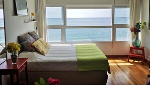 Een slaapkamer met een groot raam met uitzicht op zee, een bed met een groene deken, een nachtkastje met een vaas, en een klein krukje.