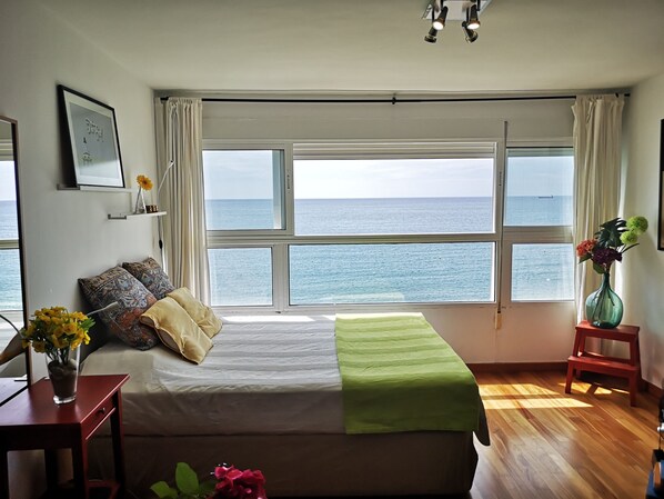 Une chambre avec une grande fenêtre donnant sur la mer, un lit recouvert d’une couverture verte, une table de chevet avec un vase et un petit tabouret.
