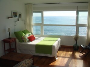Une chambre à coucher avec un lit, une table de chevet, une chaise, une fenêtre donnant sur la mer et un vase rempli de fleurs.