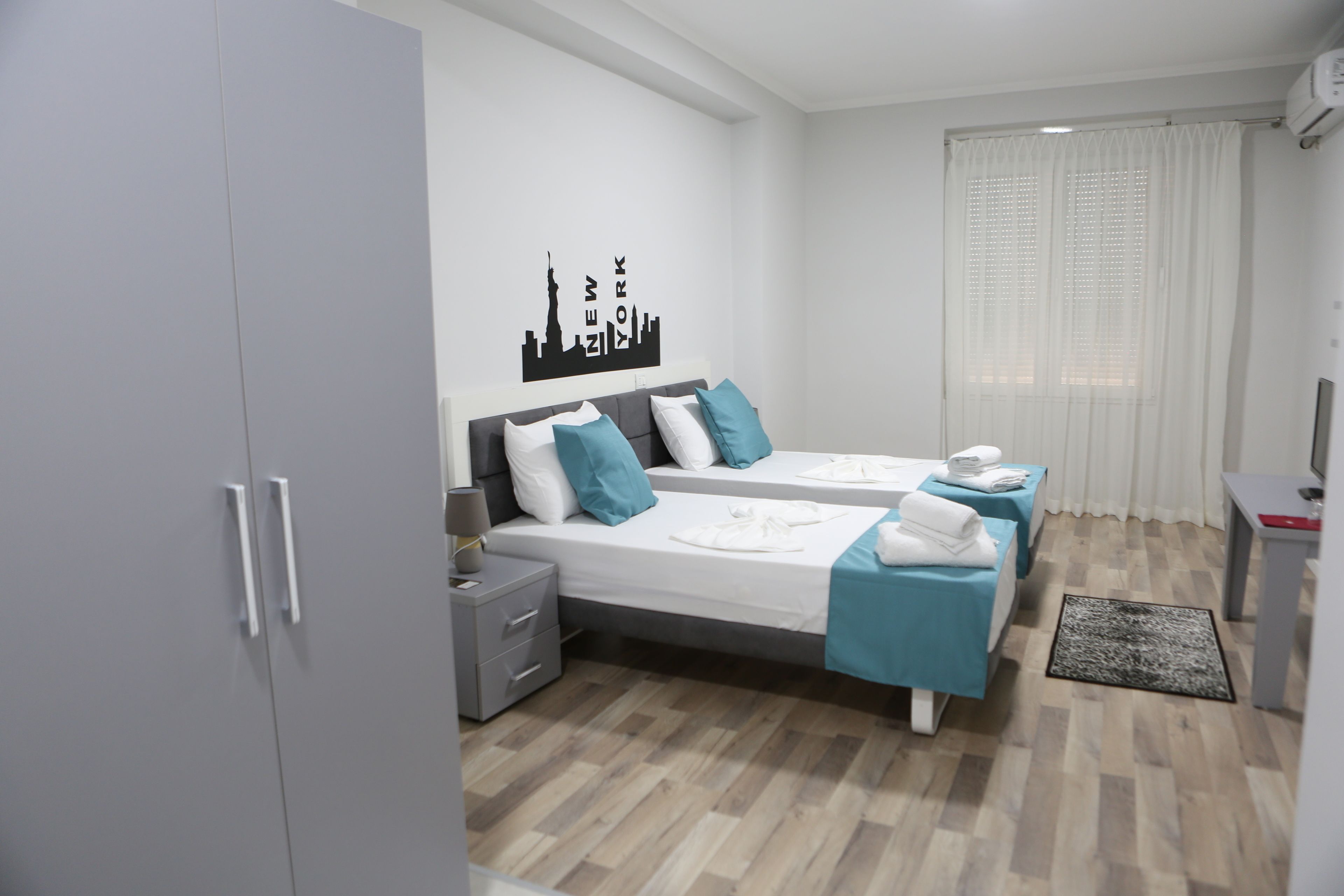 A modern hotel room with two beds, a city skyline wall art, a desk, and a window with curtains.