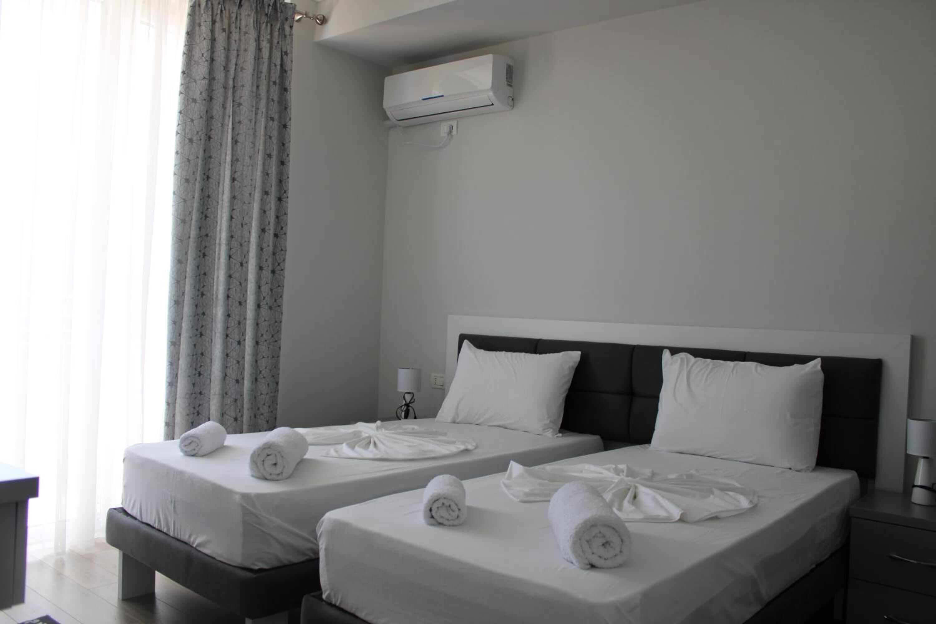 A hotel room with two beds, each with white linens and towels, a bedside table, a lamp, and an air conditioning unit on the wall.