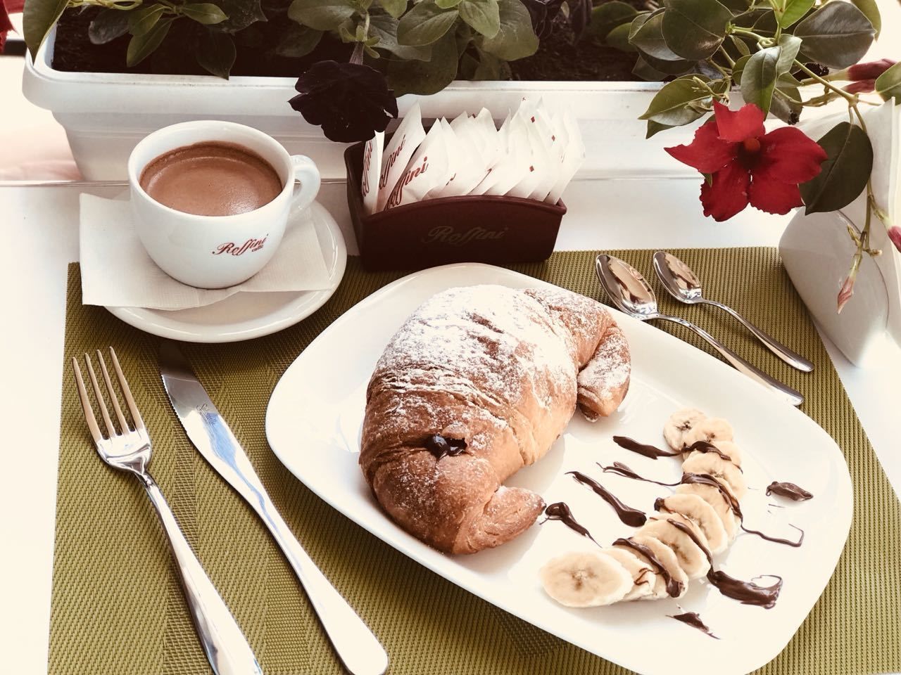 A croissant on a plate with banana slices and chocolate sauce, a cup of coffee, and utensils on a table with a placemat.