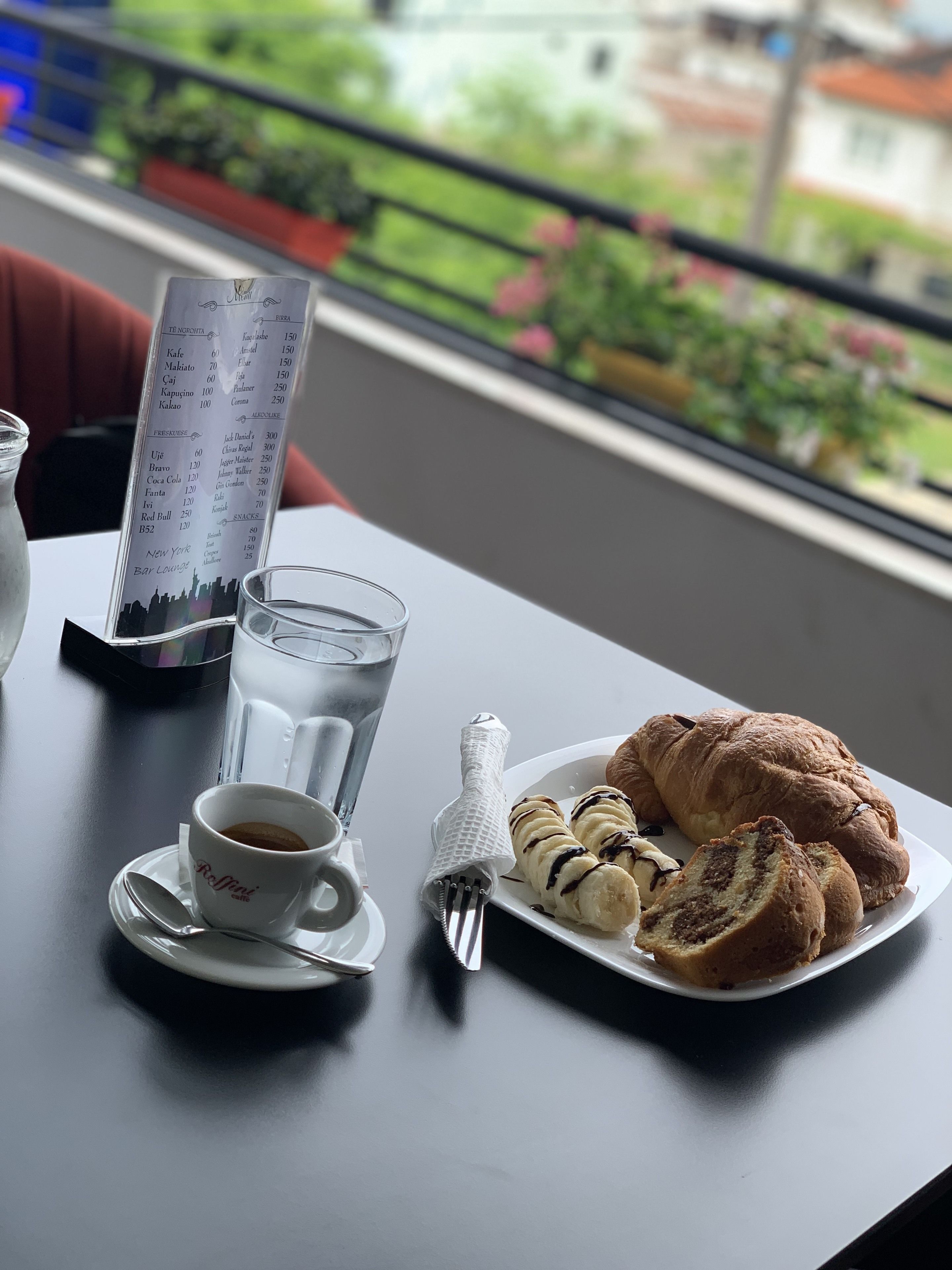 Una mesa con un menú, un vaso de agua, una taza de café, un croissant y un plato con rodajas de plátano y goteado de chocolate.