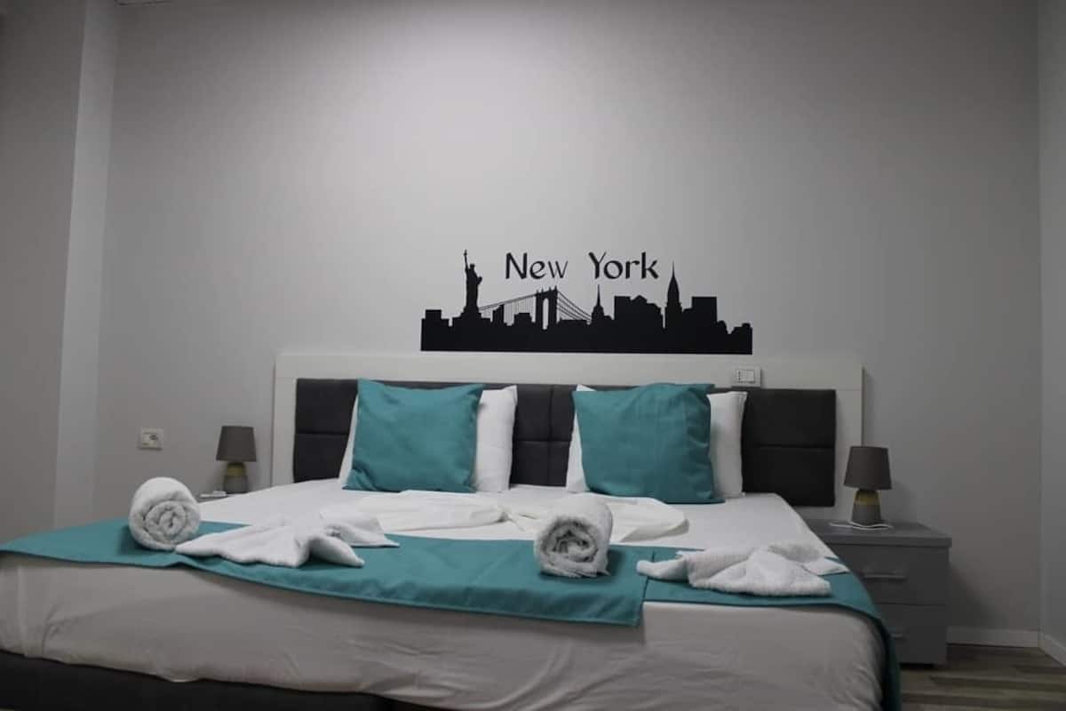 Een hotelkamer met een bed, blauwgroene beddengoed, een hoofdbord met een silhouet van de skyline van New York, twee nachtkastjes met lampen, en een in de muur bevestigde lichtschakelaar.