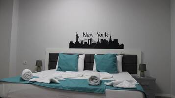 A hotel room with a bed, teal bedding, a headboard with a New York skyline silhouette, two bedside tables with lamps, and a wall-mounted light switch.