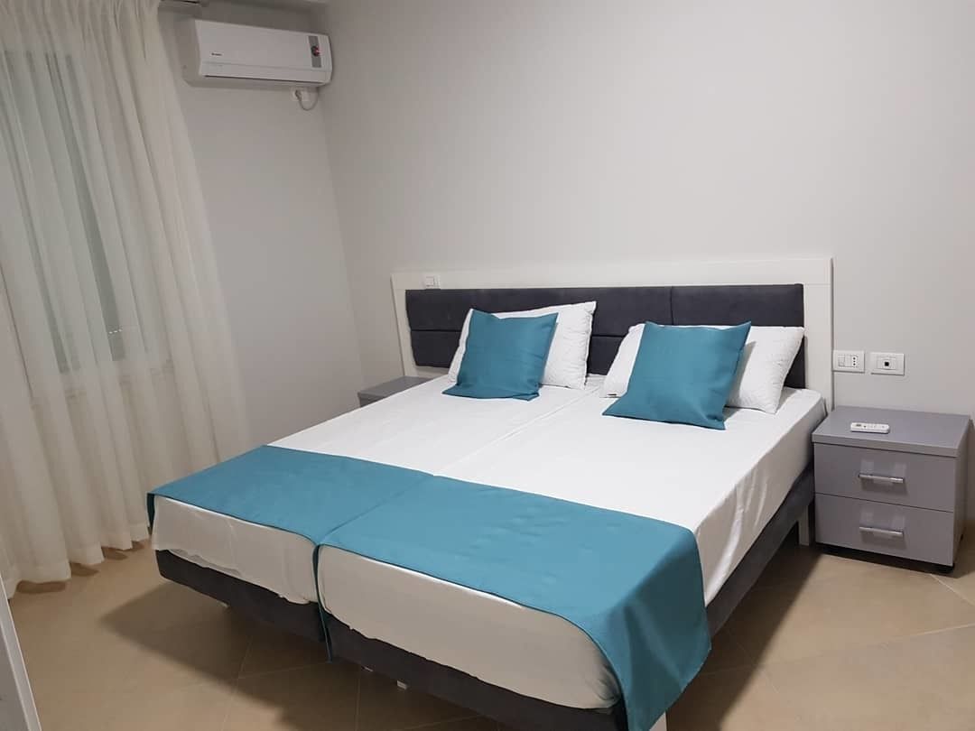 Ein Doppelbett mit weißer und blauer Bettdecke, zwei blaue Kissen, ein dunkles Kopfteil, ein Nachttisch mit Schubladen und eine Klimaanlage an der Wand.