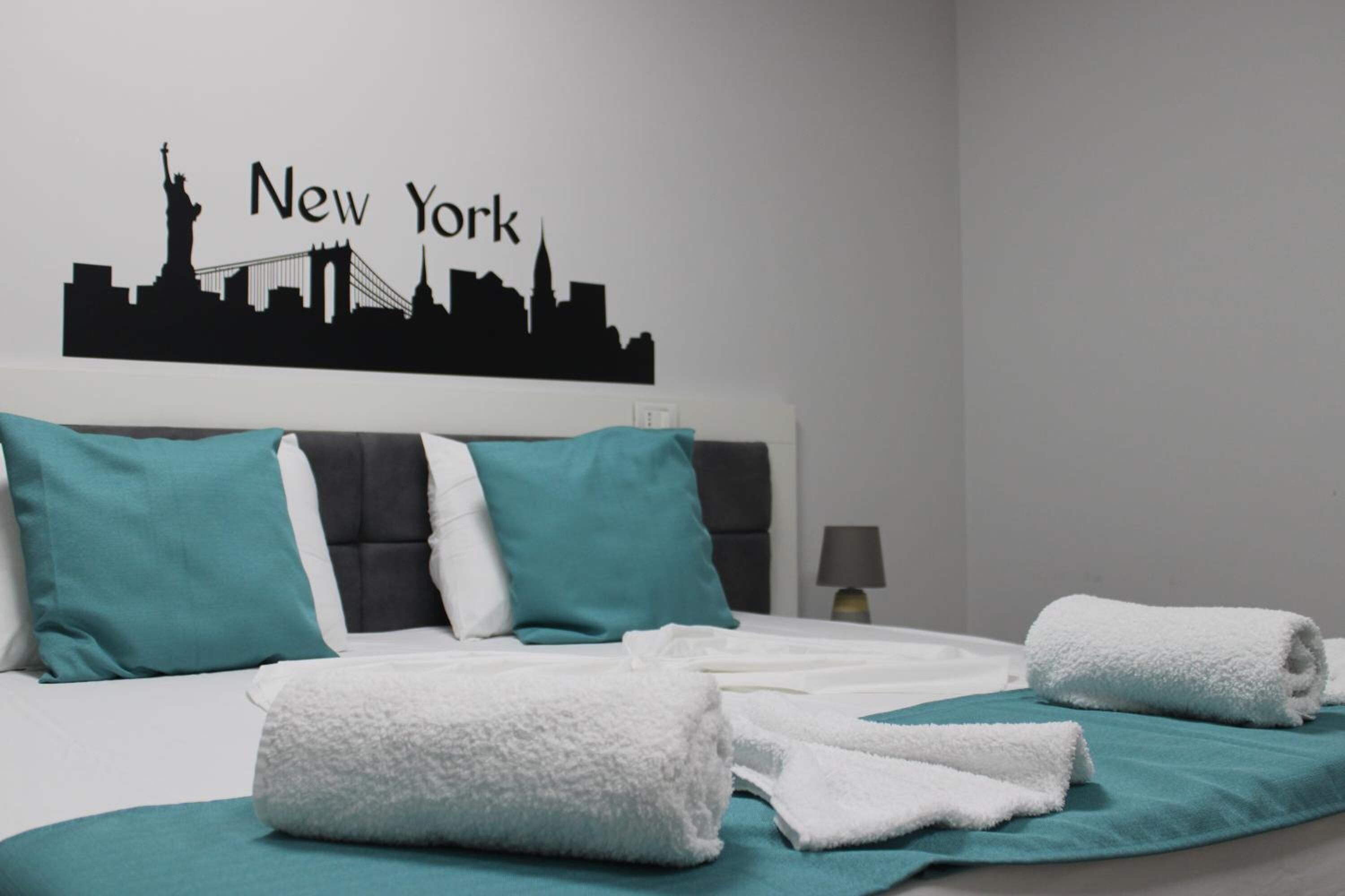 A hotel room with a bed, teal and white pillows, white towels, and a wall decal of New York skyline.