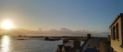 A coastal view with buildings, a clear sky, and the sun setting over the water.