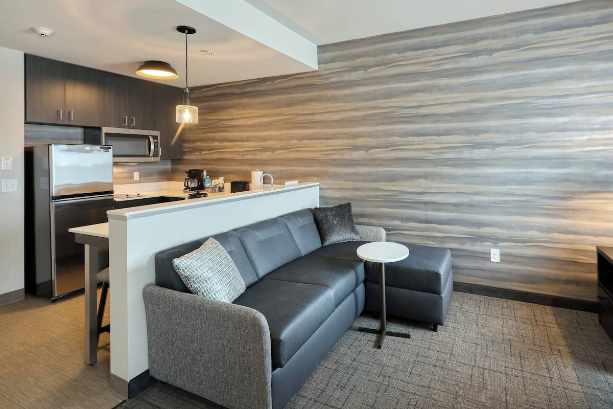 A modern kitchen with a built-in microwave and refrigerator, a gray sofa with a small side table, and a textured wall behind the seating area.