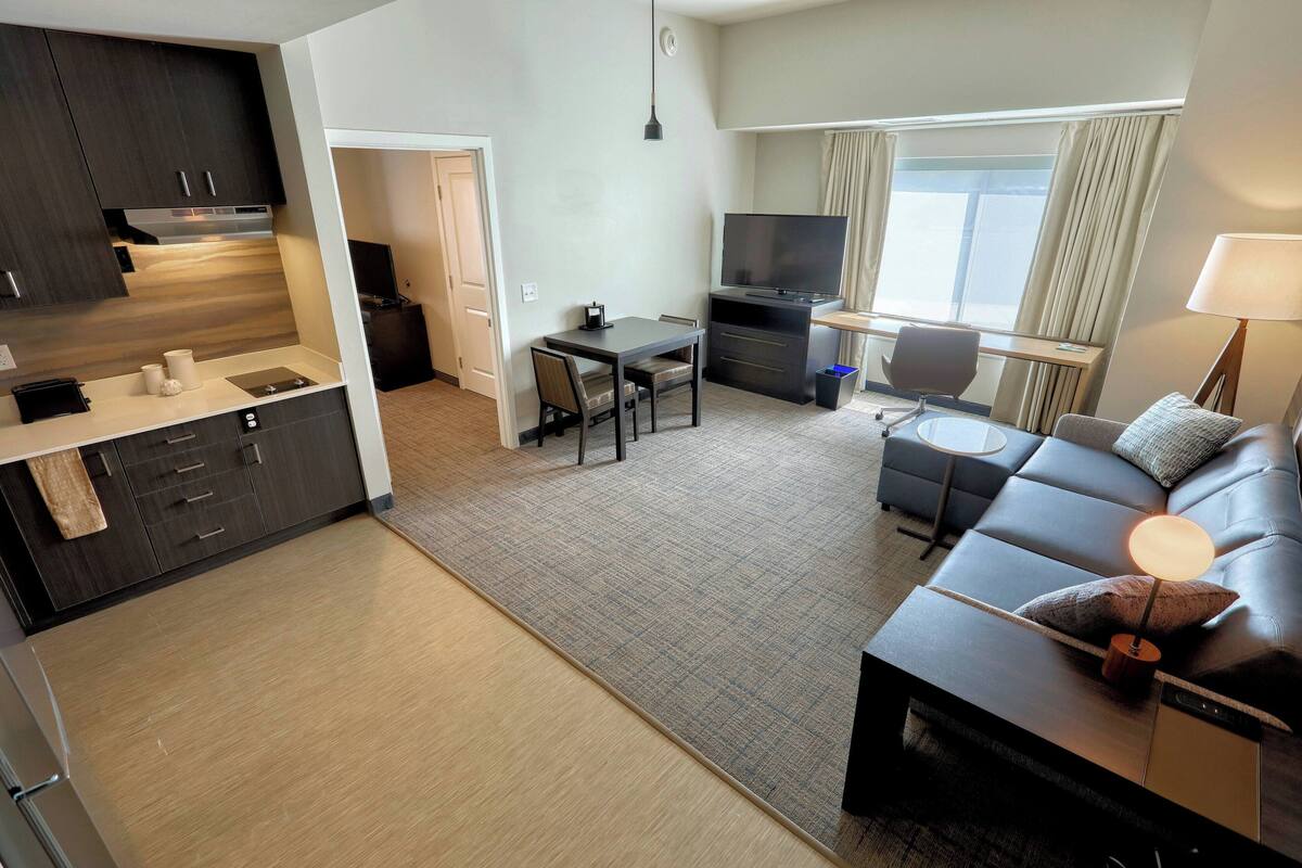 A modern hotel room with a kitchenette, a dining area, a living room with a sofa and a TV, and a large window with curtains.