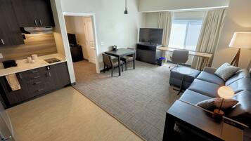Una habitación de hotel moderna con kitchenette, zona de comedor, sala de estar con sofá y televisor, y un ventanal con cortinas.