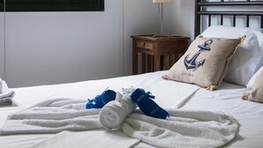 Ein ordentlich bezogenes Bett mit aufgerollten Handtüchern und einem blauen Handtuch, ein Fenster mit Jalousien, eine Nachttischlampe und ein dekoratives Kopfkissen mit Anker-Motiv.