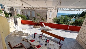 Une terrasse extérieure avec une table dressée pour le repas, entourée de fauteuils confortables et offrant une vue sur la mer et les montagnes.