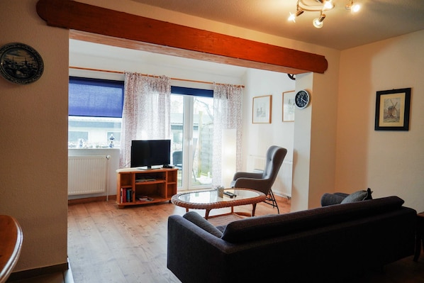 A living room with a sofa, a glass coffee table, a television on a stand, and a chair. There is a window with curtains, a clock on the wall, and a framed picture.