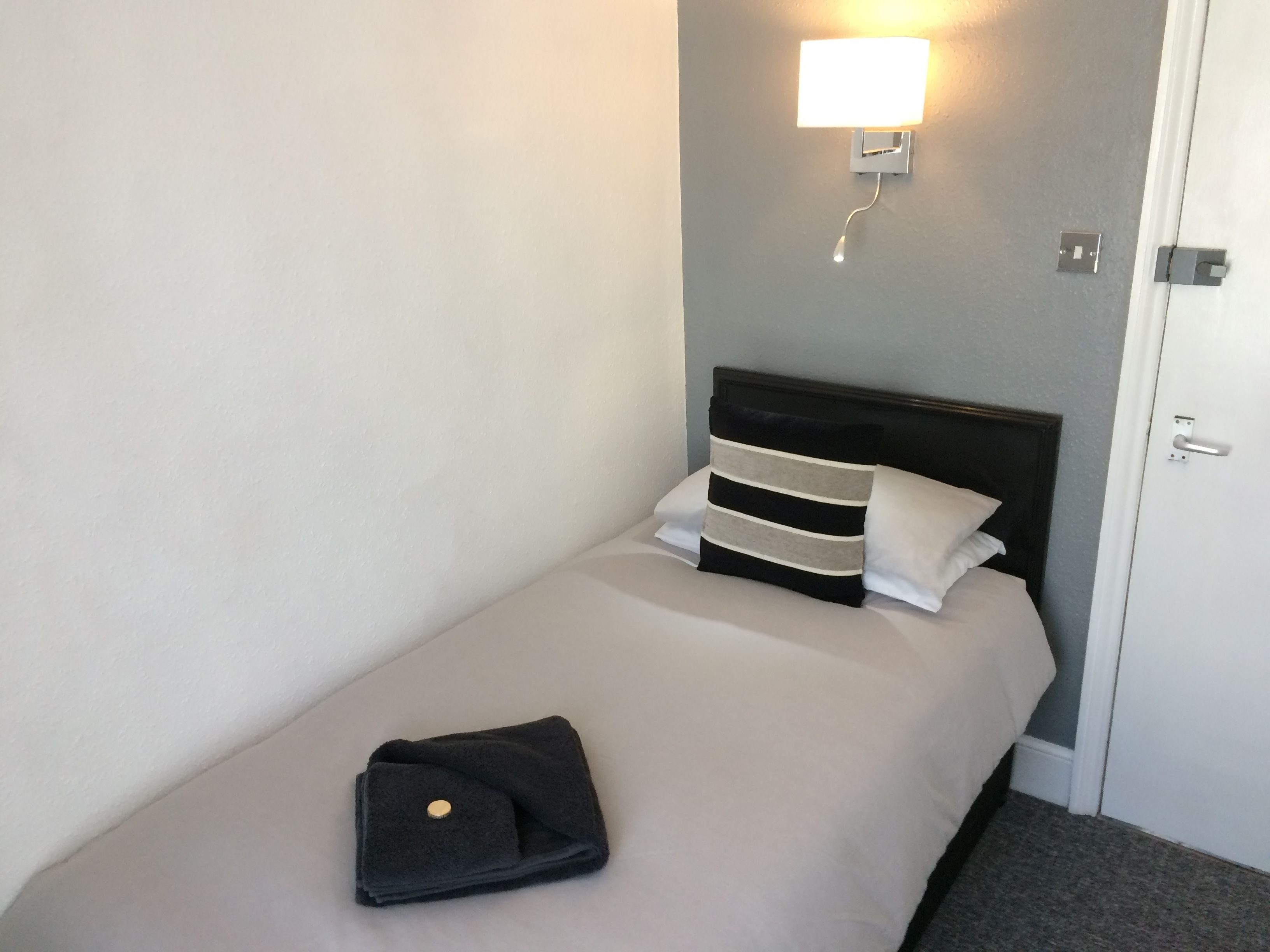 A single bed with a black and white striped pillow, a folded black towel, and a wall-mounted light fixture.