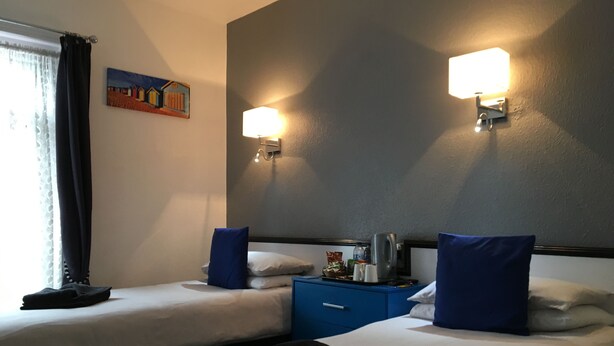 A hotel room with two beds, a nightstand, a lamp, and a painting on the wall. - Apollo Guest House (Hastings)