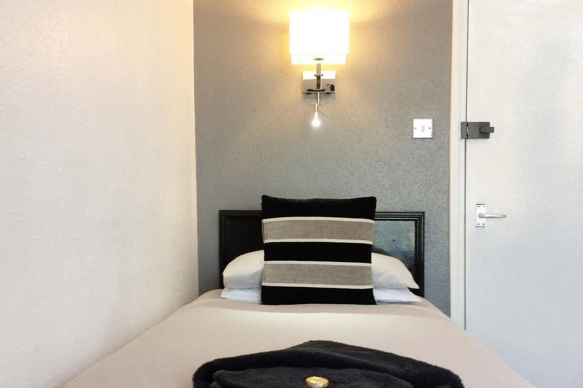 Una cama bien hecha con un almohadón a rayas, una toalla negra doblada y una lámpara de pared.