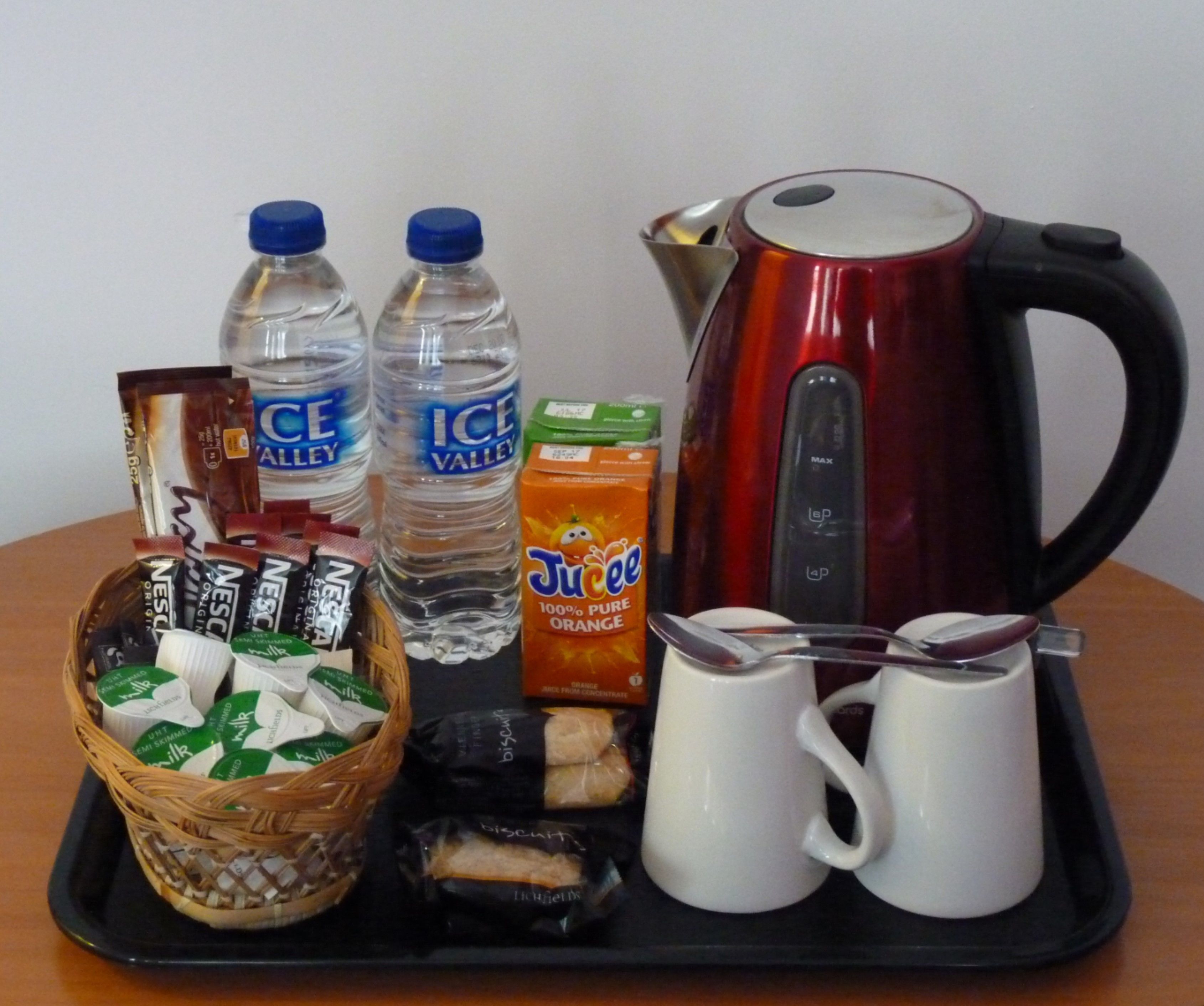 Una bandeja con una tetera, dos botellas de agua, un cartón de jugo, una cesta de galletas y dos tazas de café.