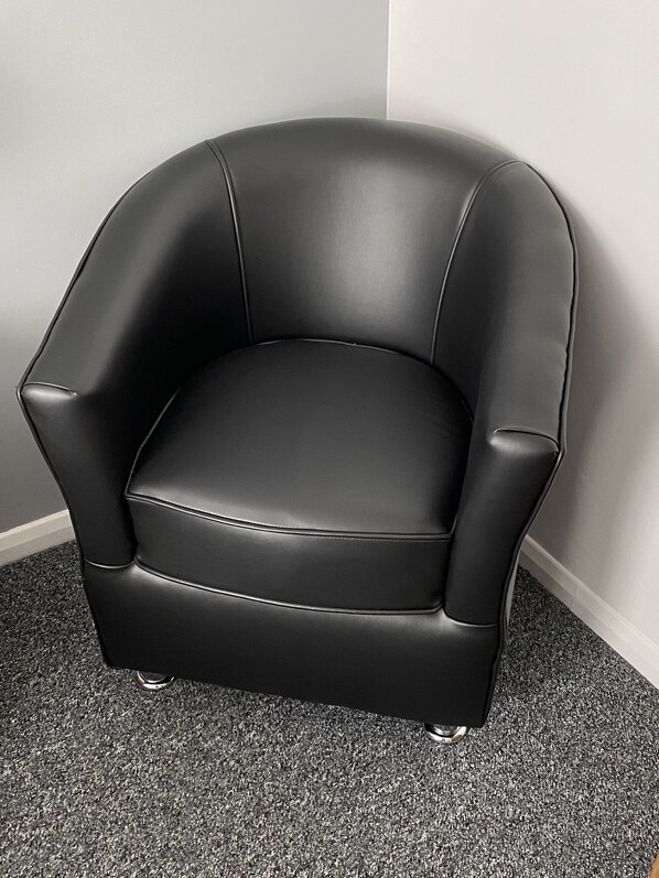 A black leather armchair with a cushioned seat and backrest, positioned against a white wall on a carpeted floor.