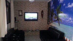 Una sala de estar con televisión instalada en una pared de azulejos, un estante en la pared y un mural tropical de gran tamaño.