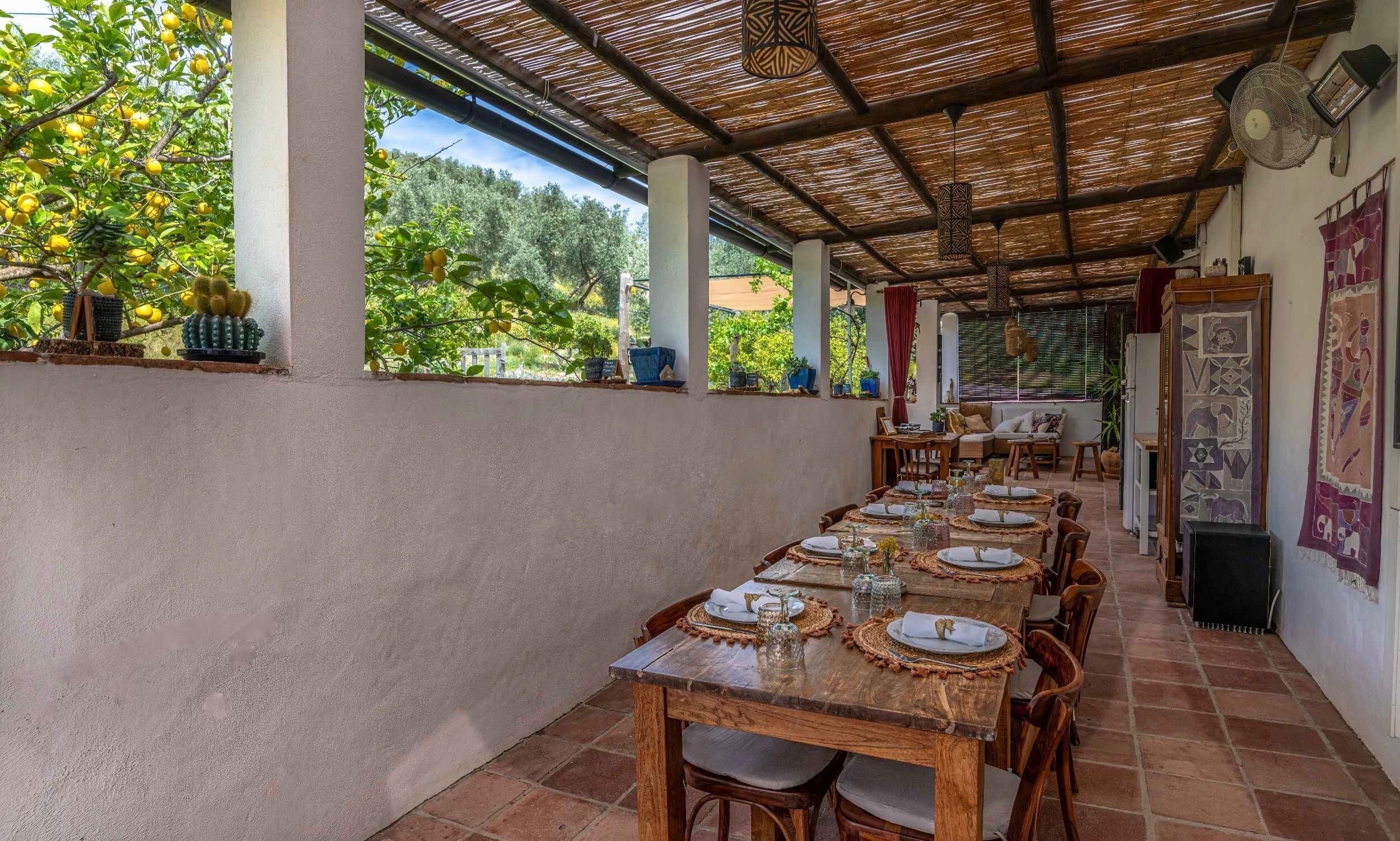 Un espace extérieur abrité, aménagé pour les repas, avec des tables dressées, entouré de plantes en pot et offrant une vue sur la verdure.
