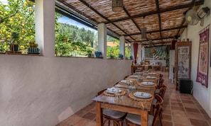 Un área de comedor al aire libre con mesas dispuestas para una comida, rodeada de plantas en macetas y vistas a follaje.