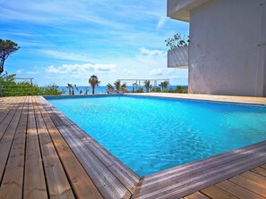 Terraza de madera alrededor de una piscina azul cristalina, con vista al océano y palmeras.