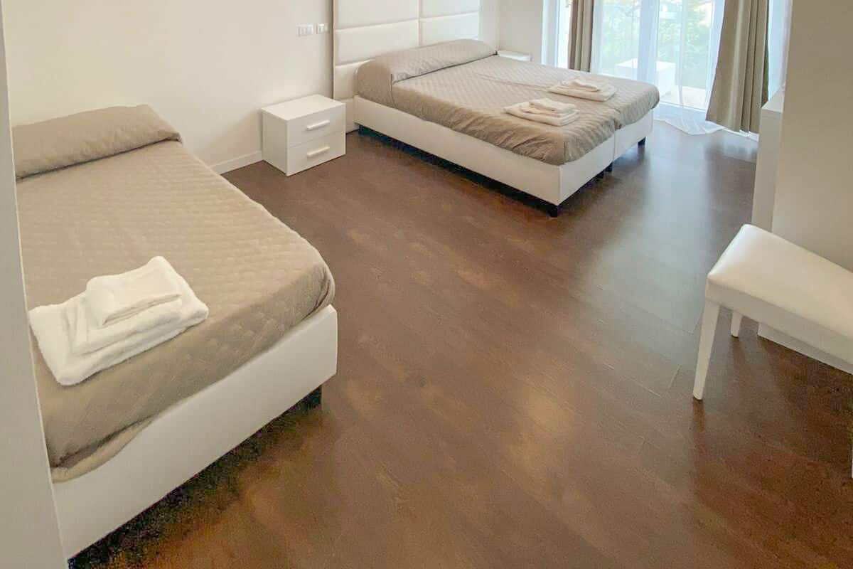 Un dormitorio moderno con una cama grande, una cama más pequeña, una mesita de noche, un banco y una ventana con cortinas.