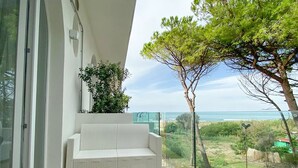 Un balcón con sofá blanco y una pequeña mesa, con vista a un paisaje costero con un árbol y el mar.