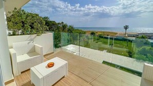 Un balcón con mobiliario de mimbre, barandilla de vidrio y vista al mar y la vegetación.
