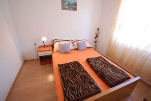 Een slaapkamer met een houten bed, twee kussens, een nachtkastje met een lamp en een raam met dunne gordijnen.