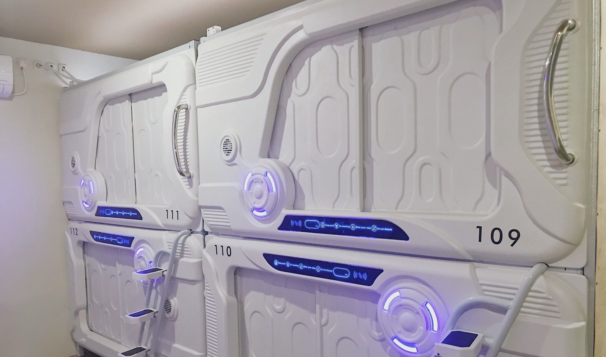 Una fila de habitaciones blancas y de estilo moderno con los números 109, 110 y 111. Cada habitación tiene un panel de control con botones iluminados y una rejilla para altavoz.