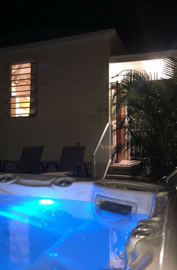 A hot tub with a blue light, a window with blinds, and a staircase leading to a balcony with palm trees.