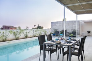 A poolside area with a table set for dining, surrounded by chairs, and a barbecue grill in the background.