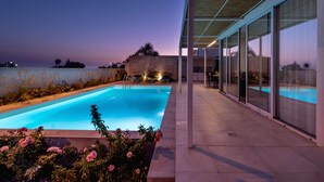 A modern outdoor swimming pool with clear blue water, surrounded by a patio area with glass walls and a wooden ceiling. - At the home of Giò, villa with pool (Marina di Ragusa)