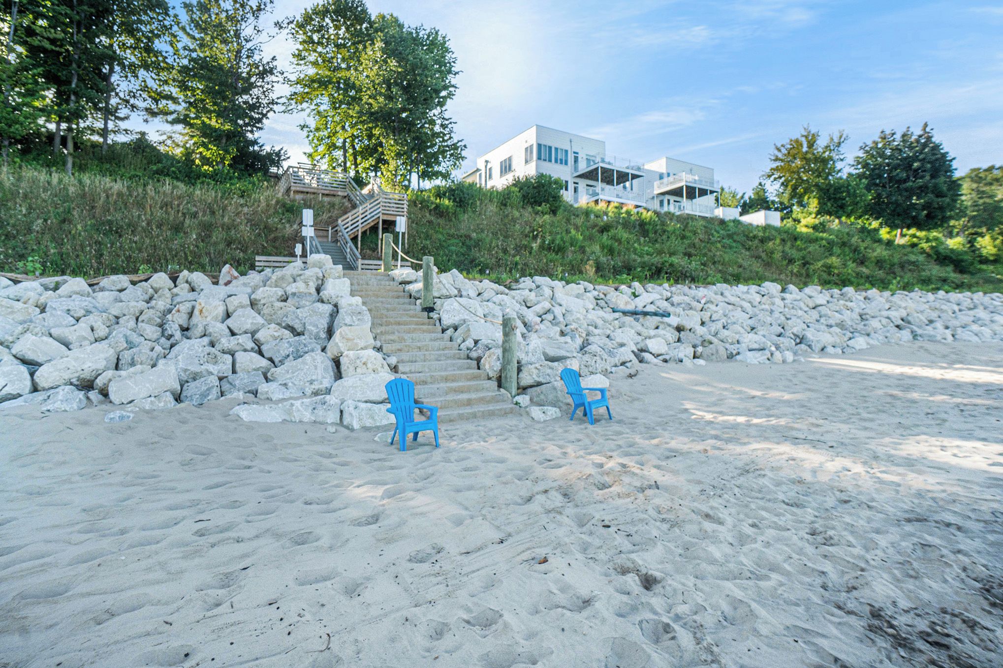A beach with two blue chairs, a stone wall, and a staircase leading to a building.