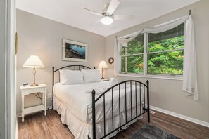 Un dormitorio con cama, lámparas de noche, ventilador de techo, ventana con cortinas e imagen enmarcada en la pared.