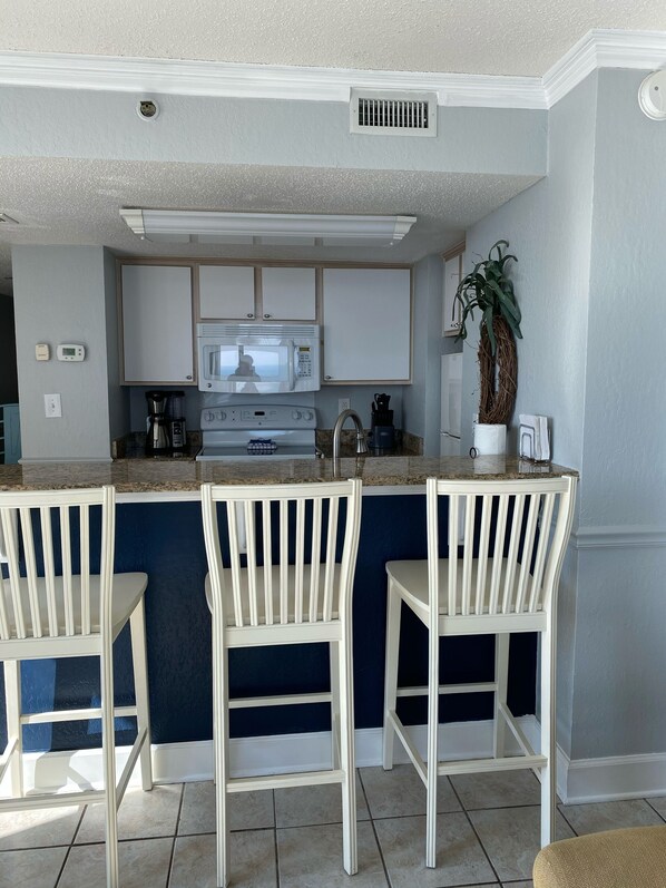 A kitchen with white cabinets, a microwave, a coffee maker, and two white bar stools. - Oceanfront W/Long Range Views, 2 bed, 2 bath Penthouse, Large Balcony, Sleeps 6 (Myrtle Beach)