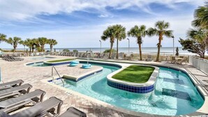 A pool area with multiple sections, including a hot tub, surrounded by palm trees and lounge chairs, overlooking a beach. - Oceanfront W/Long Range Views, 2 bed, 2 bath Penthouse, Large Balcony, Sleeps 6 (Myrtle Beach)
