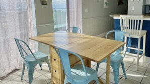 A dining area with a wooden table and four light blue chairs, a view of the sea through large windows, and a kitchen area with a white counter and blue chairs. - Oceanfront W/Long Range Views, 2 bed, 2 bath Penthouse, Large Balcony, Sleeps 6 (Myrtle Beach)